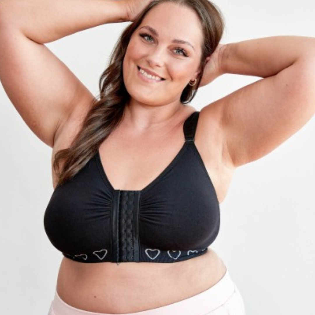 A woman wearing a post surgery comfort bra by cancer research with a front hook and eye fastening, in black with bi lateral pockets